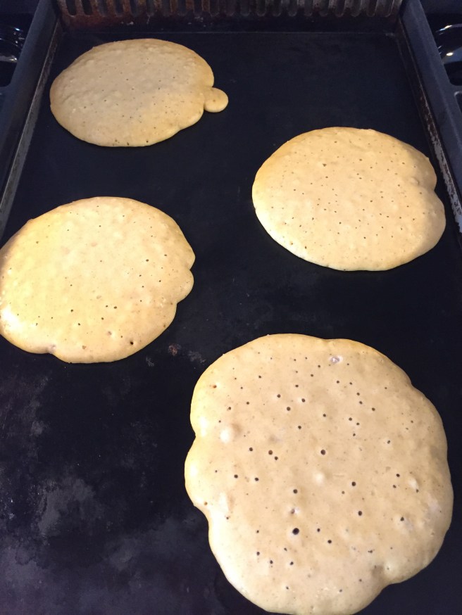 Pancakes on the griddle are dotted with popped and unpopped bubbles.