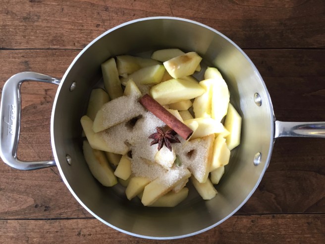 Apples with sugar, water, cinnamon stick, anise star, and cardamom pod in a saucepan.