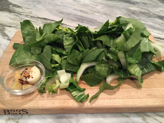 Chopped bok choy on cutting board with garlic clove, ginger, and red pepper flakes.
